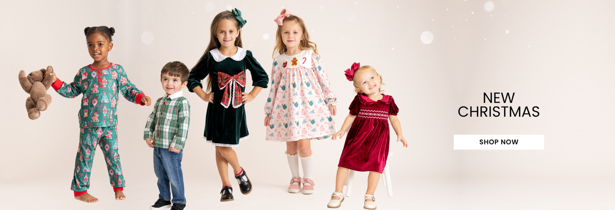 Four young children modeling holiday clothing against a light beige background with soft white bokeh lights. The children are wearing festive Christmas outfits including a green patterned pajama set, a green plaid shirt with blue jeans, a black dress with a white collar, a white dress with colorful patterns, and a dark red dress.