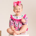 A baby wearing a pink long-sleeve romper with a white bib featuring green wreath and red bow details, along with a matching pink bow headband, sitting on a white stool against a beige background.