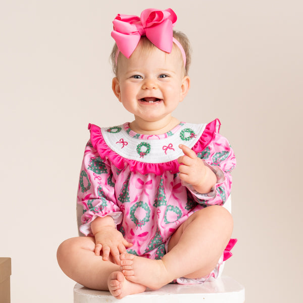 A baby wearing a pink long-sleeve romper with a white bib featuring green wreath and red bow details, along with a matching pink bow headband, sitting on a white stool against a beige background.