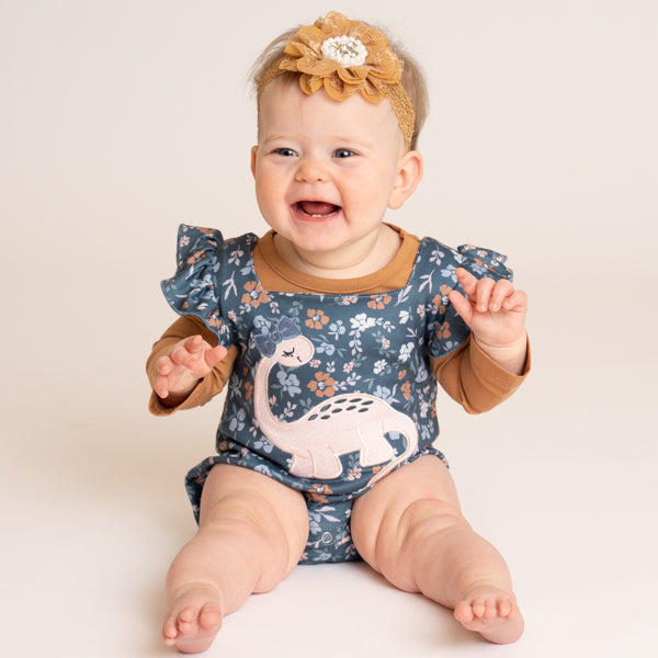 A smiling baby girl wearing a blue floral long-sleeve romper with a white dinosaur applique, brown trim, and a tan flower headband, sitting against a warm off-white background.