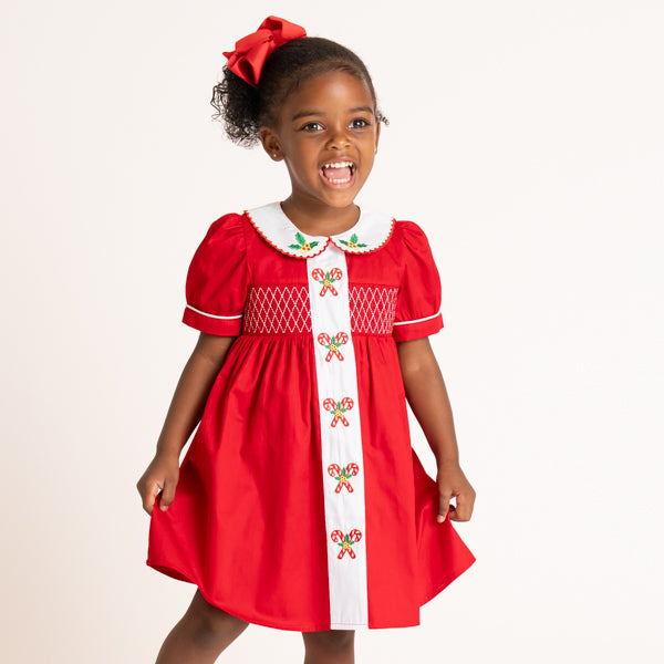 A young girl smiling and wearing a festive red dress with a white collar, white front panel, and embroidered candy cane motifs, photographed against a plain light background.