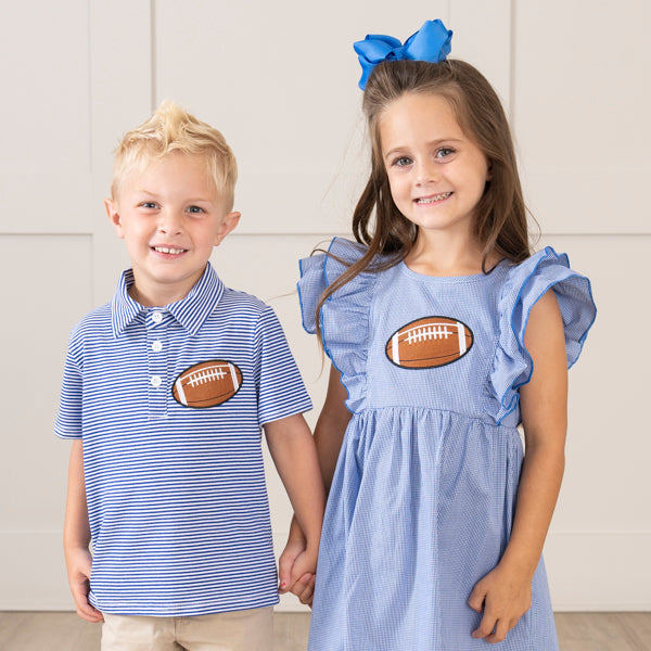 A young boy and girl standing together and holding hands. The boy is wearing a blue and white striped polo shirt with a brown football patch on the chest, and the girl is wearing a light blue ruffled dress with a matching football patch on the chest.