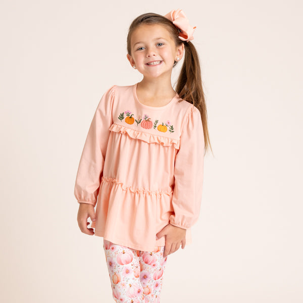 A young girl with a ponytail and a pink bow is wearing a peach long-sleeve top with pumpkin embroidery and matching floral leggings against a solid cream background.