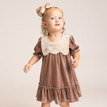 A toddler girl wearing a brown velvet dress with a cream-colored embroidered bib collar, standing against a plain beige studio background.