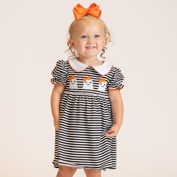 A toddler girl wearing a black and white horizontally striped dress with a white Peter Pan collar, orange trim, and embroidered smiling ghosts on the chest. She is also wearing a large orange bow in her hair, standing against a solid light beige background.