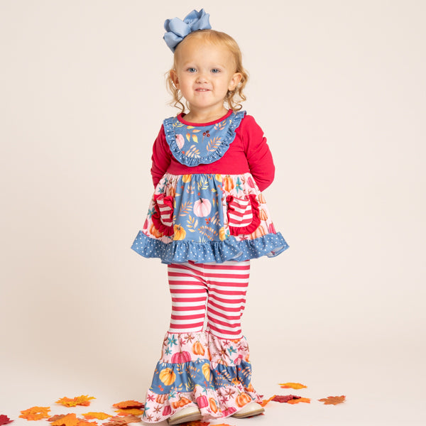 A toddler stands against a plain light beige studio background wearing a colorful autumn-themed outfit consisting of a red long-sleeve top, a blue ruffled bib-style overlay with a multicolor print, red-and-white striped leggings, and matching patterned flared pants. The child has a large blue bow in her hair and small orange leaves scattered around her feet.