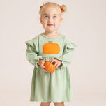 A young girl with blonde pigtails wearing a light green and white striped long-sleeve dress with an orange pumpkin applique on the chest, holding a small orange pumpkin against a solid light beige background.