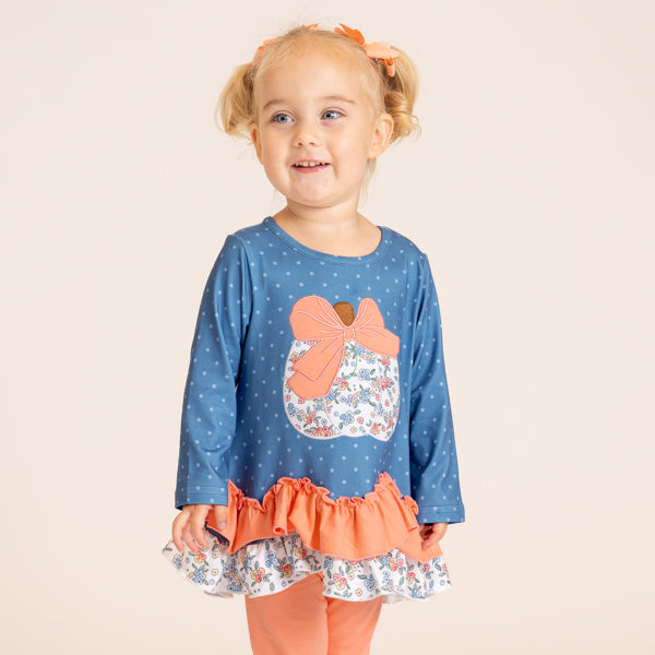 A young toddler girl with blonde pigtails wearing a blue long-sleeve top with white polka dots, a peach bow applique, and a ruffled peach and floral hem, paired with peach leggings, against a solid light beige background.
