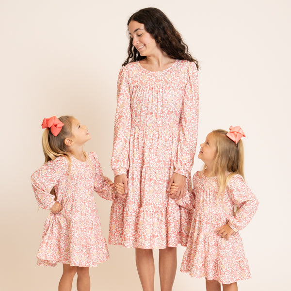 A woman and two young girls wearing matching long-sleeve floral dresses in pink and white, standing against a plain beige studio background. The woman is in the center, holding hands with the girls, who are looking up at her.