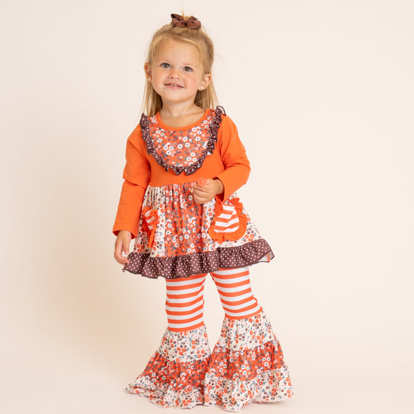 A young girl modeling a vibrant orange long-sleeve dress with a floral ruffled collar, a tiered skirt with pumpkin applique, and matching orange and white striped bell-bottom leggings, set against a plain beige studio background.