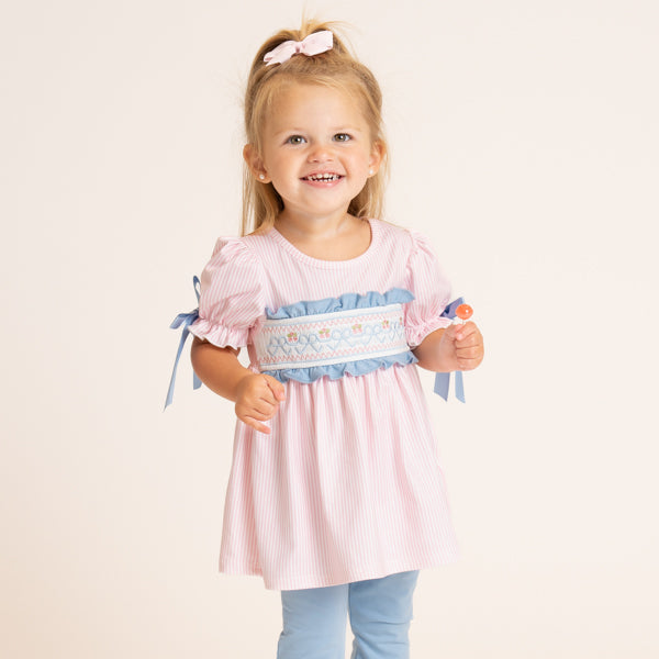 A young girl with blonde hair in a ponytail wearing a pink and white striped smocked dress with blue ribbon ties and a blue smocked waistband, paired with light blue pants, against a plain cream background.