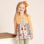 A young girl with long blonde hair and an orange bow stands smiling, wearing a long-sleeve ruffle dress with an orange and white striped top, a green button placket, and a floral skirt with pink ruffle trim. She is also wearing sage green leggings against a warm beige background.