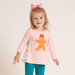 A young girl with blonde hair and a pink bow headband is wearing a light pink long-sleeve top with a gingerbread man graphic on the front, paired with teal pants, against a plain light beige background.