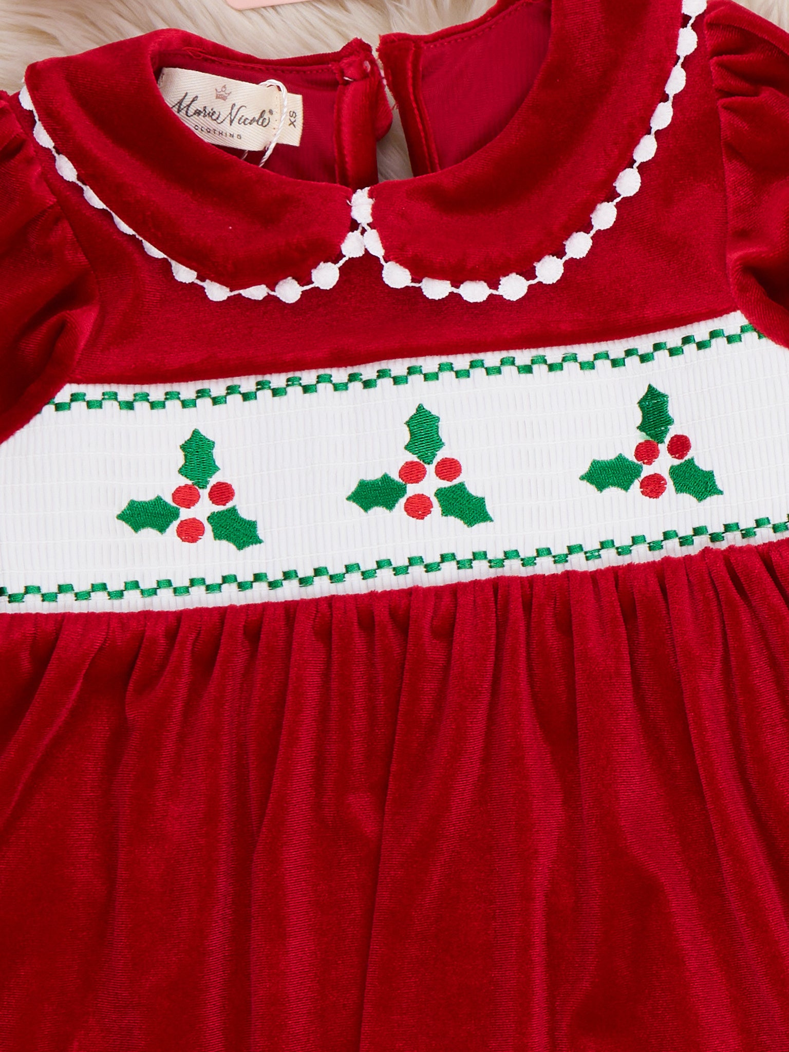 A close-up of a red velvet dress with white smocked panel featuring green and red holly embroidery and white pom pom detailing along the collar