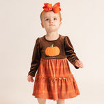A toddler girl wearing a brown velvet long-sleeve dress with an orange pumpkin applique on the chest and an orange plaid tiered skirt, paired with an orange bow headband, against a plain light beige background.