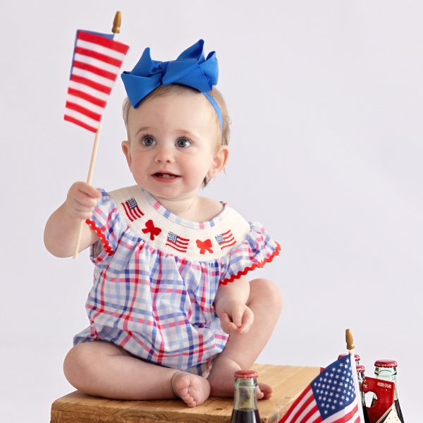 baby wears american flag romper while sitting on wooden box and holding american flag
