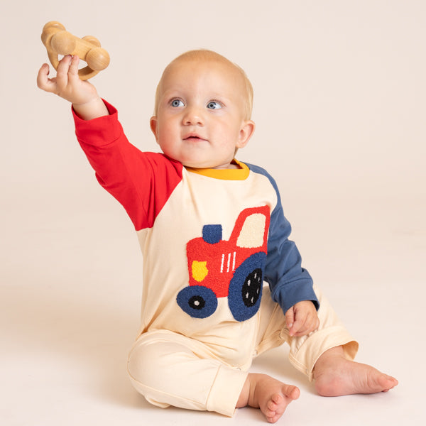 A baby sitting on a light beige studio floor, wearing a cream-colored one-piece romper with red and navy blue raglan sleeves and a red, blue, and yellow tractor applique on the chest. The baby is holding a small wooden toy in one hand.