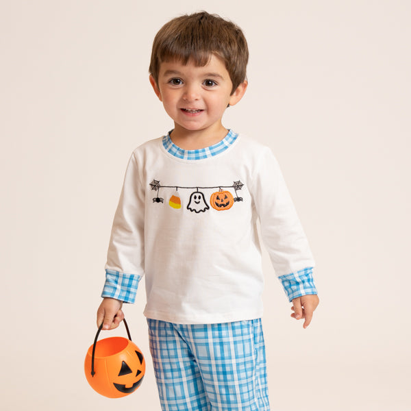 A young child wearing a white long-sleeve pajama top with a Halloween graphic and blue gingham trim, paired with matching blue gingham pajama pants, holding an orange jack-o'-lantern bucket against a light beige background.