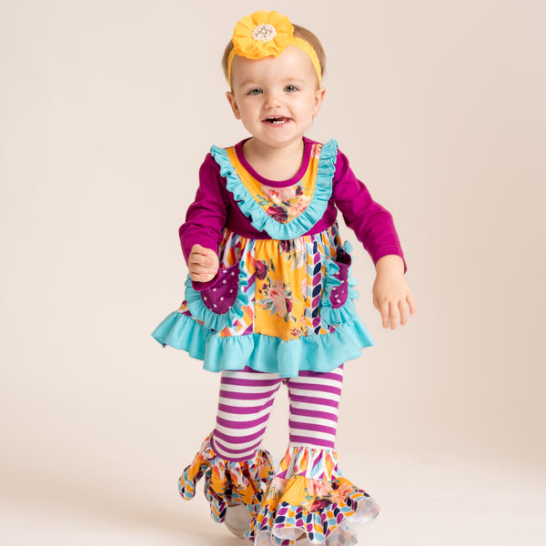 A smiling toddler stands against a plain beige studio background wearing a colorful costume-style outfit with a yellow floral headband. The outfit includes a magenta long-sleeve top, a yellow ruffled bib-style panel, turquoise ruffle trim, patterned pockets, matching patterned ruffle hems, and purple-and-white striped leggings.