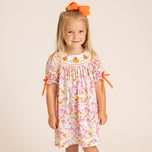 A young girl with blonde hair and an orange bow stands against a light beige background wearing a short-sleeve floral dress with orange ribbon ties on the sleeves and a smocked neckline with orange pumpkin motifs.